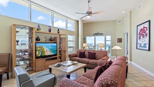 Spacious living room with a high ceiling, modern furniture, a TV showing a scenic view, and large windows providing natural light. A ceiling fan and decor items are also visible.