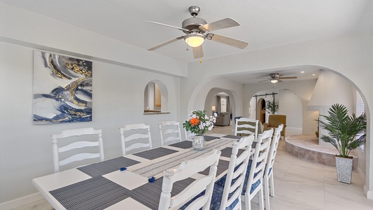 A dining room with a long table and white chairs, a ceiling fan, an abstract painting on the wall, and a view into a living area with a fireplace and another ceiling fan.