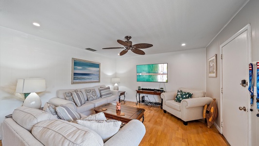 A cozy living room with a ceiling fan, three sofas, a coffee table, a TV mounted on the wall, and a painting of a beach scene. The room has wooden flooring and a closed door on the right.
