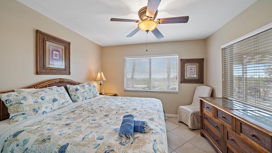 A bedroom with a large bed covered in a patterned duvet, a ceiling fan, a window with blinds, framed artwork, a bedside table with a lamp, a chair, and a dresser.