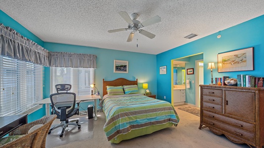 A brightly colored bedroom with teal walls features a bed with a striped comforter, a wooden dresser, a desk with a chair by the window, a ceiling fan, and an adjacent bathroom visible through an open door.