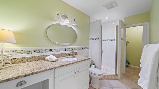 A modern bathroom with light green walls, a round mirror above a granite countertop, a white sink, a toilet, and a walk-in shower with tiled walls. Towels are hung on a rack on the right side.