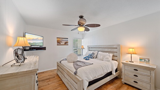 A bedroom with a double bed, two nightstands with lamps, a ceiling fan, a dresser with a TV and fan on top, and wall-mounted decorations. The room has hardwood flooring and white walls.