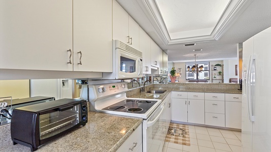 A modern kitchen with white cabinets, granite countertops, a microwave, a stovetop, a toaster oven, and a refrigerator. The backsplash has a reflective surface, and a window is visible in the background.