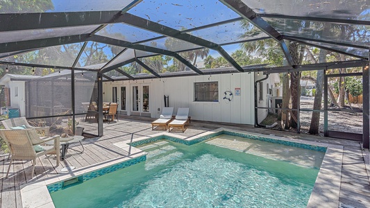 Outdoor patio area with a screened-in pool and hot tub, surrounded by lounge chairs and seating. A white building with large windows and a patio area is in the background. Trees can be seen outside the screen.