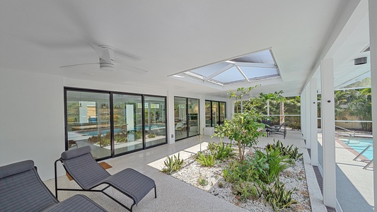 A modern indoor patio with skylights, white walls, lounge chairs, and a small garden area with plants. The space is adjacent to a pool visible through large sliding glass doors.