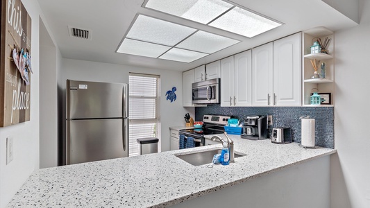 A modern kitchen featuring a stainless steel refrigerator, stove, and microwave, blue tiled backsplash, white cabinets, and a speckled countertop with a sink.