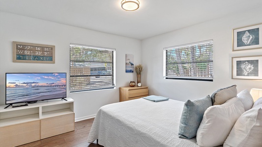 A bedroom with a white bed, light wood furniture, a TV displaying a beach scene, two windows with blinds, and beach-themed wall art.