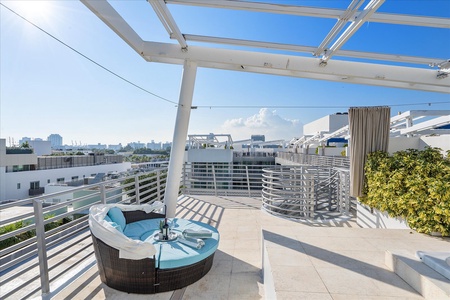 Outdoor rooftop deck with modern seating, partial shade structure, spiraling staircase, and city view. White tile flooring and a mix of greenery and building structures are visible.