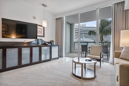 A wall-mounted TV, a seating area with a sofa and coffee table, a mini-bar, and a balcony with chairs. The balcony overlooks the pool area