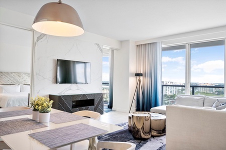 Modern living room with a dining table and TV mounted on a marble accent wall, adjacent to a bright window with city views.