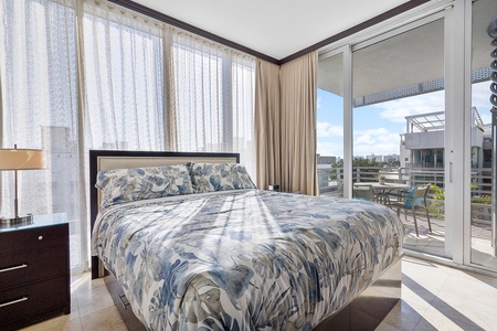 A bedroom with a floral-patterned bedspread, bedside lamp, and tall windows with sheer curtains. A glass door leads to a balcony with outdoor furniture and a view of a building.