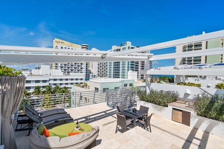 A rooftop patio with lounge chairs, a seating area, a small outdoor kitchenette, and city views on a sunny day, surrounded by modern high-rise buildings.