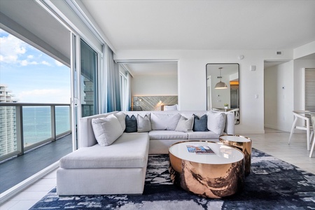 A modern open living room with a white sectional sofa that converts to a comfortable sofa bed, metallic coffee tables, and a patterned rug. Floor-to-ceiling windows reveal a balcony with an ocean view,