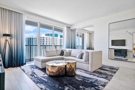 Modern living room with a large sectional sofa, round coffee tables, a floor lamp, and a large mirror. The room features floor-to-ceiling windows.