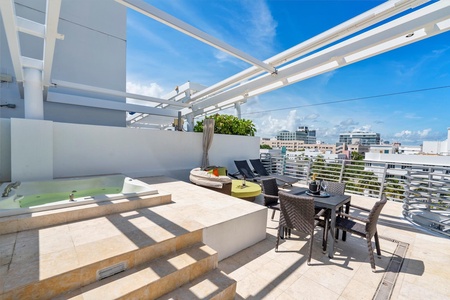 Rooftop terrace with a hot tub, lounge chairs, and a dining table with chairs under a pergola. Cityscape in the background. Sunny day with a few clouds in the sky.