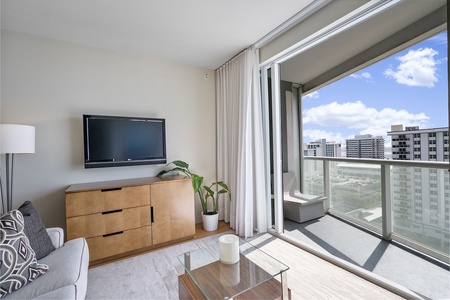 Modern living room with a wall-mounted TV, wooden dresser, sofa, and glass coffee table. A sliding glass door leads to a balcony overlooking partial ocean views and intracoastal waterway.