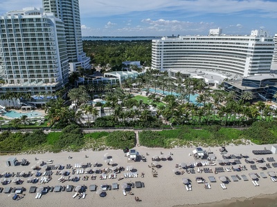 Aerial view of a beachfront hotel with several high-rise buildings, numerous lounge chairs with umbrellas set up on the sand, and lush greenery on the hotel grounds.