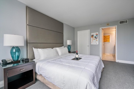 A neatly arranged hotel room with a bed, two bedside tables with lamps, a telephone, alarm and radio. An open door leads to a brightly lit bathroom.