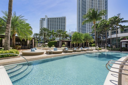 Outdoor pool area with lounge chairs, palm trees, and modern high-rise buildings in the background on a clear day.