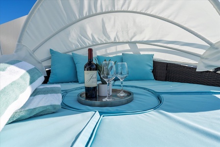 A bottle of wine and two empty wine glasses on a tray are placed on turquoise outdoor lounge furniture with pillows. A rolled-up towel is nearby, under a canopy on a sunny day.