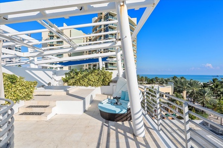 Outdoor terrace with modern white pergola, cushioned round seating, greenery, and distant tall buildings under a clear blue sky, overlooking a view of palm trees and the ocean.