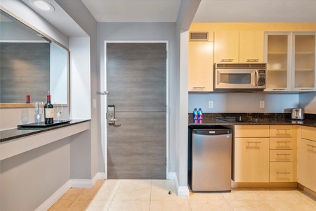 Modern kitchen with light wood cabinets, stainless steel microwave, mini fridge, and black granite countertops. A mirror and wine bottles are on a side counter near a grey door.