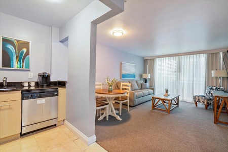 A modern kitchen and living area in a hotel suite, featuring a kitchenette with a dishwasher and coffee maker, a dining table, a sofa, and a chair, with large windows letting in natural light.