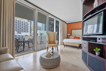 A modern hotel room with a seating area, bed, TV, and balcony access. The balcony features a table and chairs, overlooking adjacent buildings.