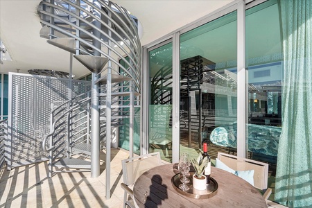 A terrace with a round table set for two, featuring wine and glasses, adjacent to glass doors and a spiral staircase, with modern architectural elements and metal railings.