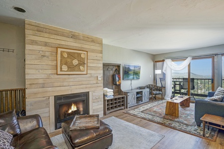 A cozy living room with a wooden accent wall, a gas fireplace, leather furniture, and a large window offering a scenic view. The room includes a TV, bookshelf, and artwork above the fireplace.