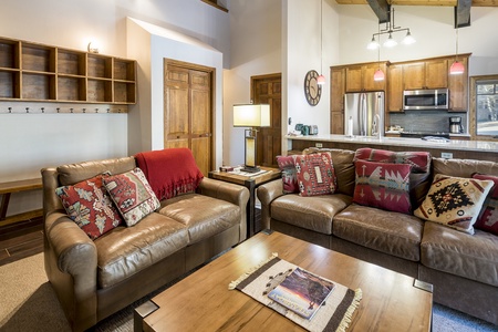 A cozy living room with leather sofas, colorful pillows, a wooden coffee table, and a view into a modern kitchen with stainless steel appliances.