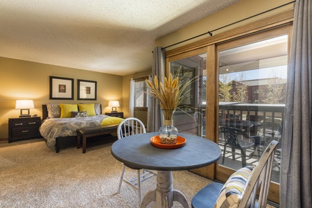 A well-lit bedroom with a small round dining table, chairs, and a glass door leading to a balcony. The bed has a patterned comforter and yellow pillows. A vase with dried wheat is on the table.