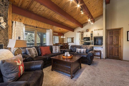 A cozy living room features dark leather sofas, a wooden coffee table, a vaulted wooden ceiling with beams, and a view into a kitchen and dining area. Natural light enters through large windows.