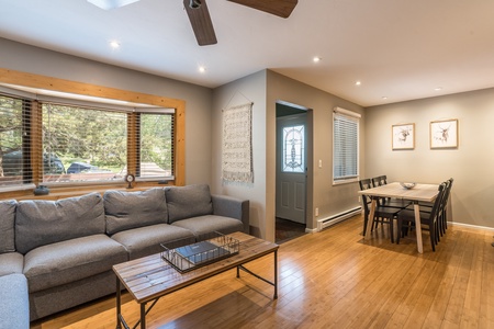 A cozy living room with a gray couch, wooden coffee table, and wall art. A dining area with a table and chairs is visible in the background. Sunlight enters through a large window with wooden trim.