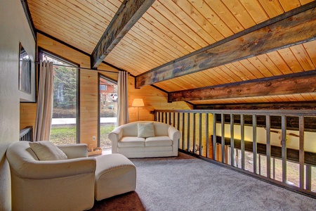 A cozy loft area featuring a pitched wooden ceiling with exposed beams, two white armchairs, a small table, a lamp, and large windows with views of the outdoors.