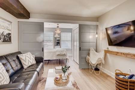 A living room with a black leather couch, a wooden coffee table, and a wall-mounted TV. The room opens into a bedroom with a bed and two large windows. A wicker basket and throw blanket are in the corner.