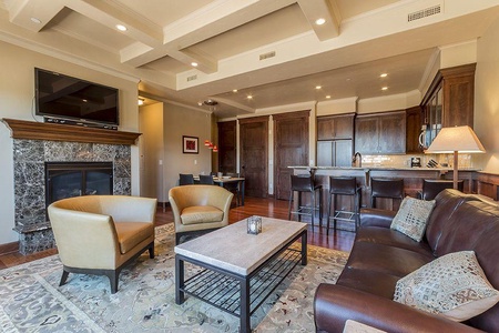 A cozy living area with a marble fireplace, two beige chairs, a brown leather sofa, a coffee table, and a TV. The background includes a kitchen with a bar counter and wooden cabinetry.