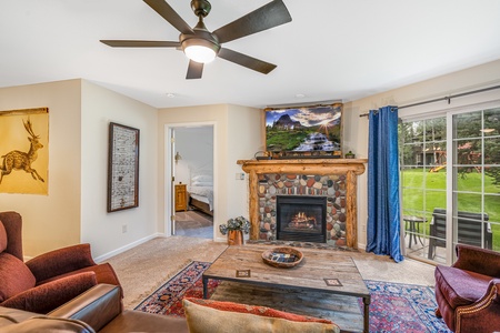 Spacious living room with fireplace, ceiling fan, and large windows. A red sofa, a coffee table, and a framed wall painting are visible. A bedroom is partially seen through an open door. A backyard is outside.