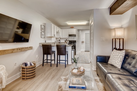 A modern living room features a black leather couch, a wall-mounted TV, a minimalist kitchen island with two stools, and a woven basket with blankets on a wooden floor.