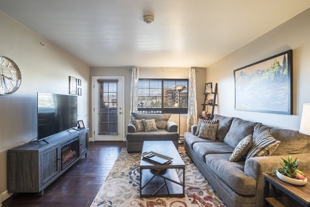 A cozy living room featuring a grey couch, an armchair, a flat-screen TV on a media console, a coffee table, and a wall clock. Natural light enters through a window and a glass door leading outside.