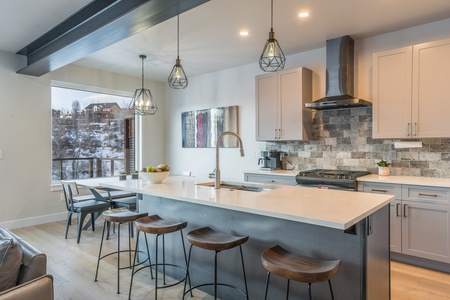 Modern kitchen with a central island featuring four bar stools, pendant lights, a mounted range hood, and a dining area by a large window with a view of snow-covered trees.