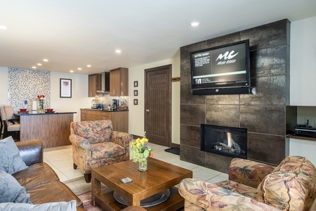 A cozy living room with a fireplace, TV, and floral armchairs. The space includes a wooden coffee table, a brown leather sofa, and an open kitchen area in the background. A vase with flowers on the table.
