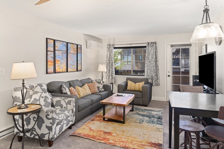 A living room with a grey couch, armchair, wooden coffee table, and rug. There is a TV on a table, a dining table with chairs, artwork on the wall, and a window with curtains.