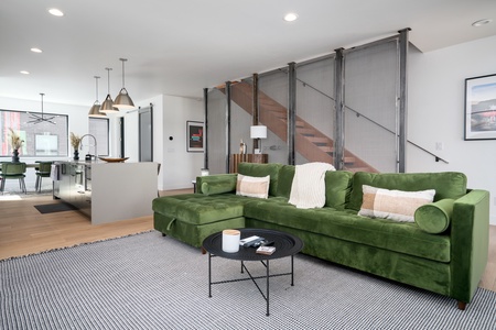 Modern living room with a large green sofa, a black coffee table on a gray rug, a staircase with metal mesh railing, and a view of an open kitchen and dining area in the background.