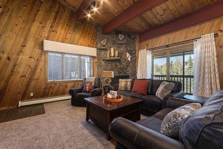 A cozy living room with wooden walls, a large stone fireplace, dark leather sofas, a wooden coffee table, and windows with white curtains. There are decorative pillows and a tray on the table.