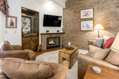 A cozy living room with a lit fireplace, brick wall, two brown armchairs, a beige sofa, a wooden chest as a coffee table, and mounted TV. Decor includes framed pictures and a lampshade.