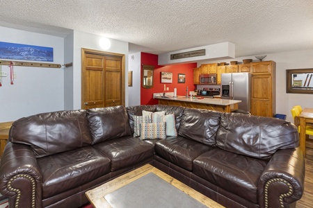 A living room with a brown leather sectional sofa, a wooden coffee table, and a view of a kitchen area with wooden cabinets and stainless steel appliances in the background.