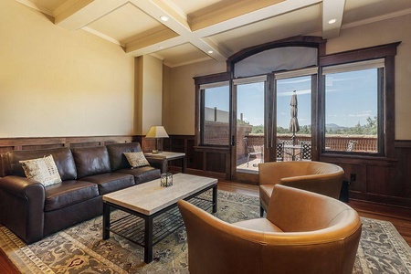 A cozy living room with a brown leather couch, two tan chairs, a coffee table, and a large window overlooking an outdoor patio area. The room features wooden paneling and a beige ceiling with beams.