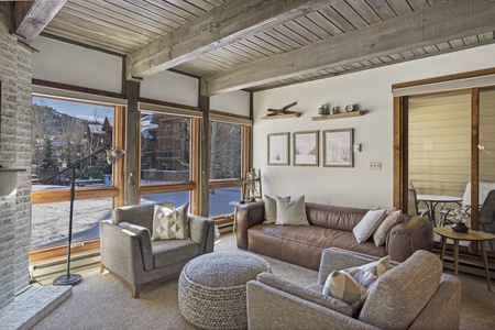 A cozy living room with wooden beams, large windows overlooking a snowy landscape, a brown leather sofa, gray armchairs, and framed wall art. A sliding door leads to a small balcony.